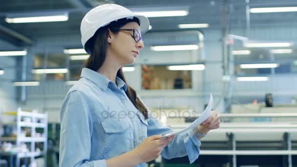 Beautiful Brunette Industrial Engineer Wearing Hard Hat Studies Blueprints and Documents while Walking Through Factory. — Stock Video