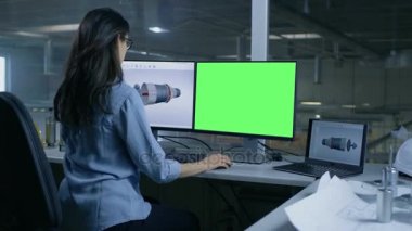 Profesyonel kadın mühendis tasarımlar 3d türbin / motoru büyük bir sanayi şirketi için onun ikinci monitör Mock-up yeşil ekran bilgisayar gösterir. Ofis pencere büyük fabrika dışında görülür.