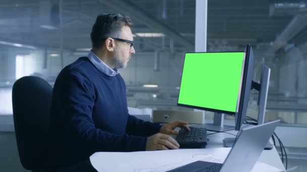 Chef-Ingenieur, der an seinem Personencomputer an technischen Projekten arbeitet. Das zweite Display zeigt eine grüne Bildschirm-Attrappe. Aus dem Bürofenster sieht man eine große Fabrik. — Stockvideo