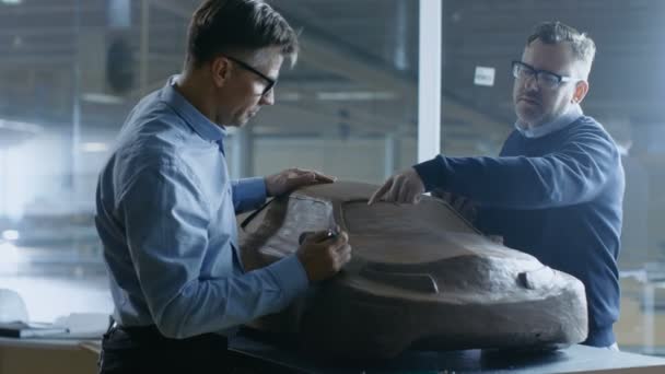 Two Male Automotive Designers Working on a Clay Model of New Generation Electric Car Future Design. One Holds Tablet Computer, Other Sculpts with Clay with Rake/Wire. — Stock Video