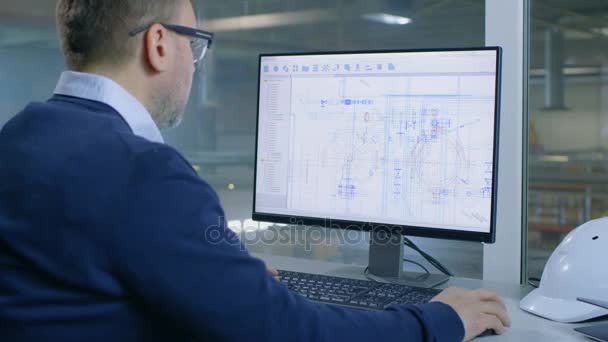 Der männliche Wirtschaftsingenieur arbeitet an seinem Computer mit technologischen Bauplänen. Das Innere der Fabrik ist von ihrem Bürofenster aus zu sehen. — Stockvideo