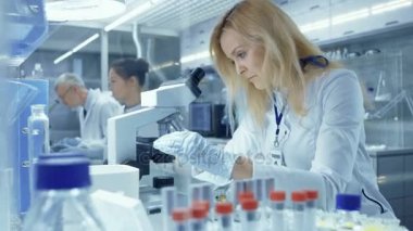 Kadın araştırma bilim adamı mikroskop altında biyolojik örnekleri inceliyor. O ve arkadaşları büyük bir Modern laboratuarda çalışmak.