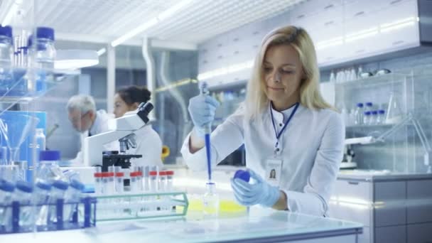 Ученая-женщина использует микропипетки для заполнения пробирки в большой современной лаборатории. На заднем плане Ученые работают . — стоковое видео