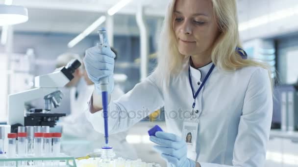 O cientista de pesquisa fêmea usa tubos de teste de enchimento de micropipeta em um grande laboratório moderno. No fundo, os cientistas estão trabalhando . — Vídeo de Stock