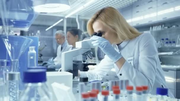 Forscherin betrachtet biologische Proben unter dem Mikroskop. Sie und ihre Kollegen arbeiten in einem großen modernen Labor / medizinischen Zentrum. — Stockvideo