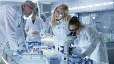 Tıbbi Araştırma Ekibi Yeni Nesil Hastalık Tedavisi üzerinde çalışıyor. Mikroskop, Test Tüpleri, Mikroplar ve Yazı Analiz Sonuçları kullanıyorlar. Laboratuvar meşgul, parlak ve modern görünüyor..