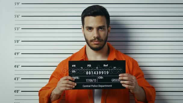 In una stazione di polizia arrestato uomo ottenere lato, Front-View Mug Shot. Indossa una tuta arancione da prigioniero e tiene Placard. Grafico di altezza sullo sfondo . — Video Stock