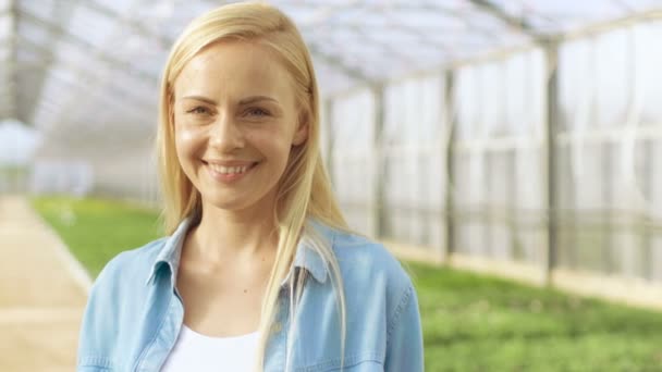 An einem sonnigen Tag steht die schöne blonde Gärtnerin lächelnd in einem Gewächshaus voller bunter Blumen. — Stockvideo