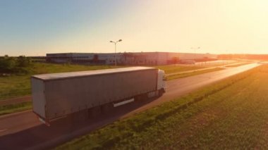 Beyaz yarı kamyon kargo Trailer karayolu üzerinde hareket ile hava görünümünü. Arka plan depolar ve endüstriyel yükleme yapılar görülmektedir. Günbatımı.