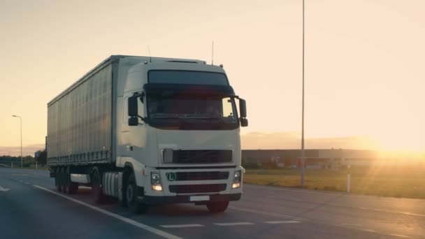 Seguimento Tiro de um semi-caminhão com reboque de carga se movendo em uma estrada. Caminhão branco dirige através da área do armazém industrial nas primeiras horas da manhã . — Vídeo de Stock