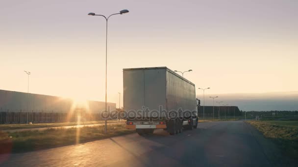 Následná střela návěsu kamionu s poplatky na dálnici. Bílý vůz projede průmyslové manipulační plochy na prázdné silnici s slunce zářící na pozadí.