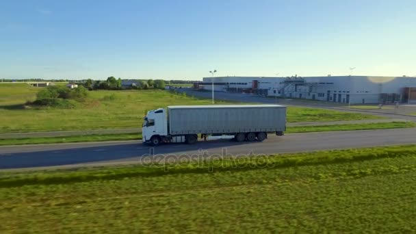 Luchtfoto zijaanzicht van witte Semi vrachtwagen met lading aanhangwagen verplaatsen op de snelweg. In de achtergrond magazijnen en landelijke omgeving, mooie zonnige dag. — Stockvideo