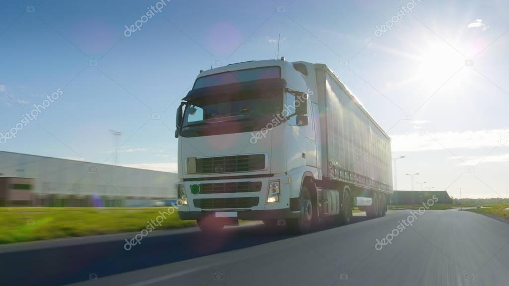 Big White Semi Truck with Cargo Trailer Moves on the Industrial Area Empty Road With Sun Shining in the Background. Blur motion.