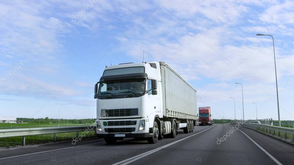 Speeding White Semi Truck with Cargo Trailer Drives on the Highway. Truck is First in the Column of Heavy Vehicles, Sun is Shining.