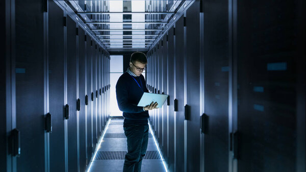 Male IT Engineer Works on a Laptop in front of Server Cabinet at