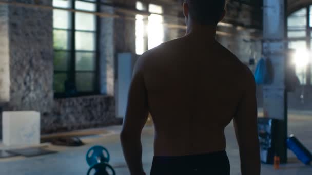 Suivi Prise de vue d'un homme sans chemise musclé entrant dans une salle de sport au ralenti. Il est confiant, le bâtiment est industriel et hardcore, divers équipements de fitness / bodybuilding couchés sur le sol . — Video