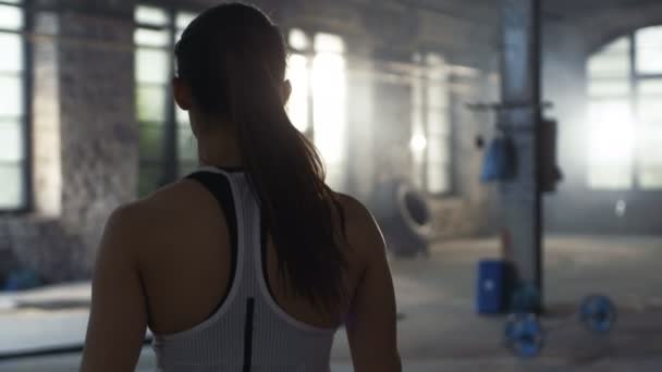 Seguimiento de Shot of Athletic Beautiful Woman Entrando en el gimnasio en cámara lenta. Ella es confiada y tira de su cola de caballo, Edificio es Industrial y Hardcore, Varios Cross Fitness / Culturismo Equipo acostado en el suelo . — Vídeos de Stock