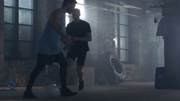 Dvě svalové fotbalistů hraje s míčem v tělocvičně budovy průmyslového. Oni mají zábavu, různé posilovací zařízení je ležící na podlaze. — Stock video