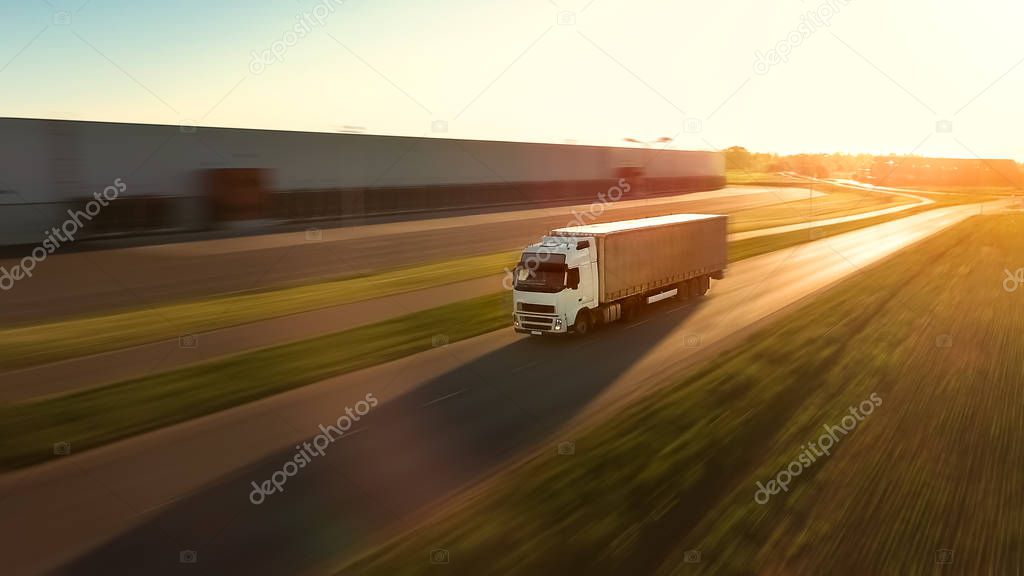 Aerial View of White Semi Truck with  Cargo Trailer Moving on the Highway. In the Background Warehouses and Rural Area, Sun is Setting. Motion Blur.