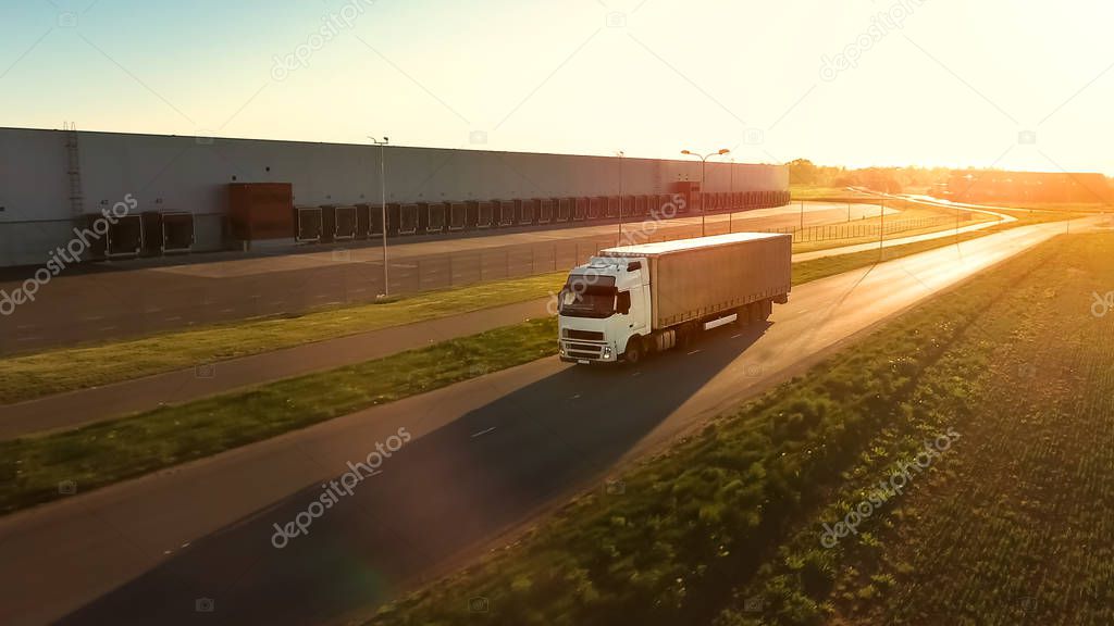 Aerial View of White Semi Truck with  Cargo Trailer Moving on the Highway. In the Background Warehouses and Rural Area, Sun is Setting.