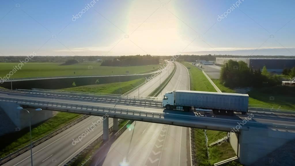 Aerial Shot of the White Truck with Semi Trailer Moving on the Highway. In the Background Warehouses and Rural Area, Sun is Setting.