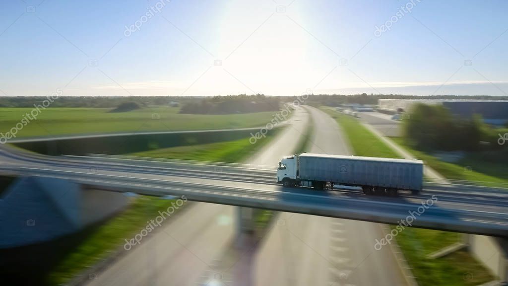 Aerial Shot of the White Truck with Semi Trailer Moving on the Highway. In the Background Warehouses and Rural Area, Sun is Setting. Motion Blur.