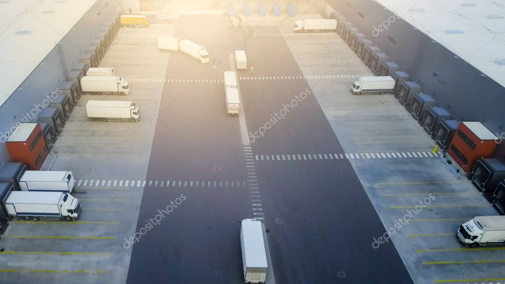 Aerial Shot of Truck with Attached Semi Trailer Leaving Industrial Warehouse/ Storage Building/ Loading Area where Many Trucks Are Loading/ Unloading Merchandise.