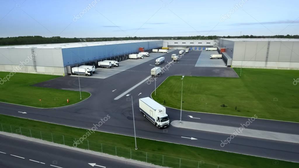 Aerial Shot of Truck with Attached Semi Trailer Leaving Industrial Warehouse/ Storage Building/ Loading Area where Many Trucks Are Loading/ Unloading Merchandise.