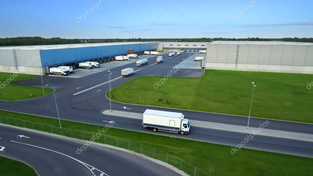 Aerial Shot of Truck with Attached Semi Trailer Leaving Industrial Warehouse/ Storage Building/ Loading Area where Many Trucks Are Loading/ Unloading Merchandise.