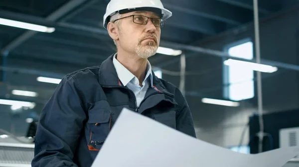 Ingeniero senior en hardhat está de pie en una fábrica y mirando — Foto de Stock