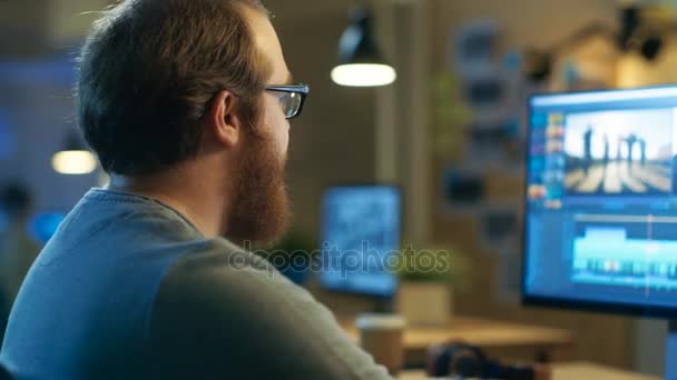 Der männliche Videofilmer bearbeitet und schneidet Material und Ton auf seinem PC mit zwei Displays. sein Büro ist ein modernes und kreatives Loft-Studio. — Stockvideo