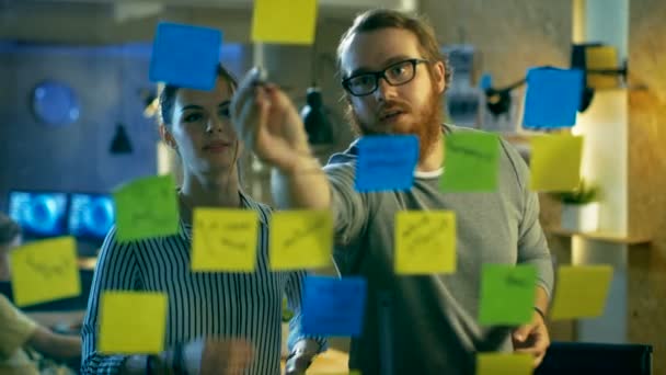 Male and Female Designers Discuss and Outline They're Project with Sticky Notes on a Glass Wall. They're Young and Creative People Who Work in a Stylish Loft Space. — Stock Video