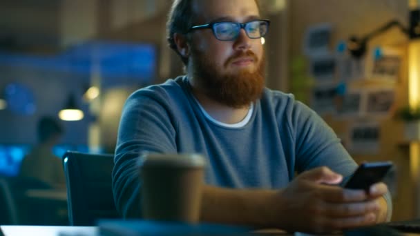 Jonge, bebaarde Man typen bericht op zijn Smartphone. Hij zit aan het Bureau en de glimlach. — Stockvideo