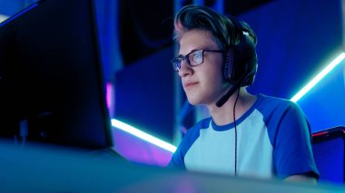 Teenage Boy Gamer Plays in Competitive Video Game on a eSports Tournament/ Internet Cafe. He Wears Glasses and Headphones with Microphone. clipart