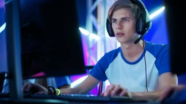 Professional Boy Gamer Plays in Video Game on a eSports Tournament/ in Internet Cafe. He Wears  Headphones and Speaks Commands into Microphone. clipart