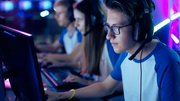 O jogador profissional do menino joga no jogo vídeo em um torneio de eSports / café do Internet. Ele usa óculos e fones de ouvido com microfone. Outras meninas e meninos jogadores jogando em segundo plano . — Fotografia de Stock