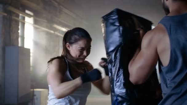 Спортивная женщина тренирует свои удары по боксерской сумке, которую держит ее партнер / тренер. Она сильная и великолепная женщина. Они тренируются в спортзале . — стоковое видео