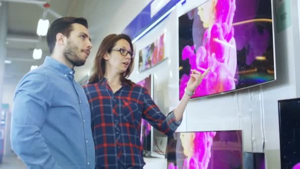 Jovem casal Compras para um novo conjunto de TV 4K na loja de eletrônicos. Estão a tentar decidir o melhor modelo, mas têm dúvidas. . — Vídeo de Stock