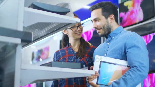 Hermosa pareja joven en la tienda de electrónica Navegación, en busca de nuevos gadgets, tabletas y cámaras de fotos / video presentados en los estantes . — Vídeos de Stock