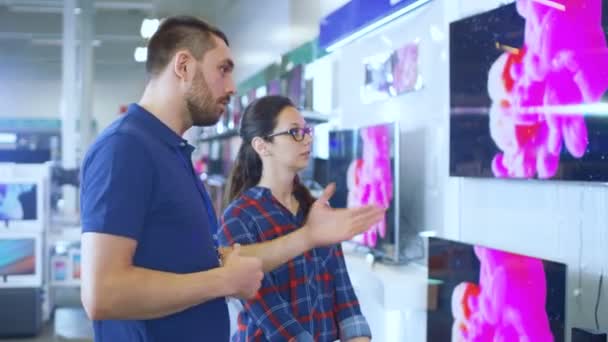 In the Electronics Store Professional Consultant Provides Expert Advice to a Young Woman Who Looks for a  New 4K UHD TV to Buy. In This Bright, Modern Store all the Latest TV Models Are Hanging on the Wall. — Stock Video