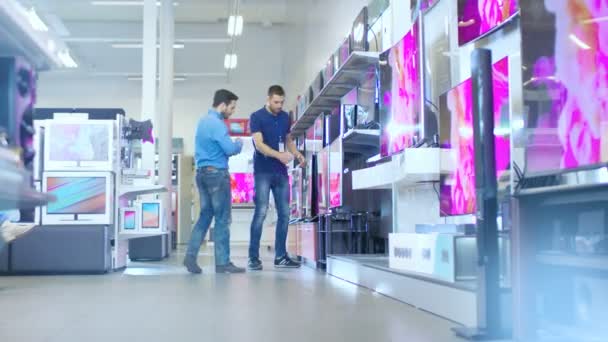 In the Electronics Store Professional Consultant Shows Latest 4K UHD TV's to a Young Man, They Talk about Specifications and What Model is Best for Young Man's Home. Store is Bright, Modern and Has all the Latest Models. — Stock Video