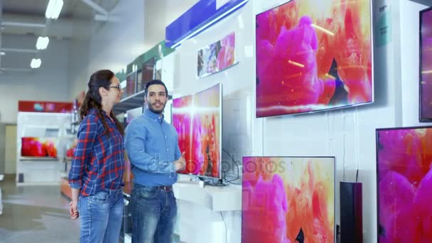 En la tienda de electrónica, el consultor profesional se acerca a la pareja joven con su asesoramiento experto sobre cuál de los nuevos modelos de televisores 4K es mejor para su hogar y presupuesto . — Vídeos de Stock