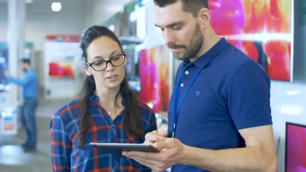 En la tienda de electrónica, el consultor profesional proporciona asesoramiento experto en las especificaciones de la computadora de la tableta para la hermosa mujer joven. La tienda es brillante, moderna y tiene todos los dispositivos más recientes . — Vídeos de Stock