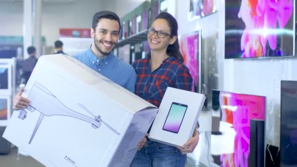 En la tienda de electrónica Happy Young Couple Poses with Newly Purchased Drone and Tablet Computer. La tienda es grande y brillante, tiene todas las últimas televisiones, cámaras y teléfonos inteligentes . — Vídeos de Stock