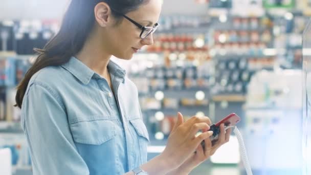 Joven hermosa mujer mira el último modelo Smartphone en la tienda de electrónica, Considera compraren ella. La tienda es brillante y moderna, llena de nuevos dispositivos . — Vídeos de Stock