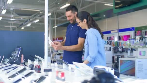 Na loja de eletrônicos lindo casal jovem olha para o computador tablet mais recente, Eles pensam em comprar um. Loja é grande, brilhante e tem todos os novos dispositivos . — Vídeo de Stock