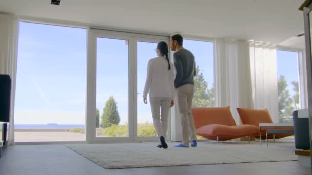 Hermosa pareja joven en su casa recién comprada, miran el mar a través del piso hasta las ventanas del techo. Su casa de lujo es brillante, moderna con interior hecho con buen gusto . — Vídeos de Stock