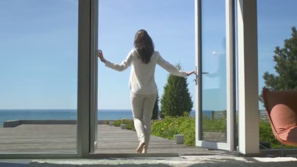 Folgende Aufnahme einer schönen Brünetten mit lockerem Haar, die die Balkontüren öffnet und auf die sonnige Terrasse mit Meerblick geht. es ist wolkenloser Morgen und sie ist wirklich glücklich. — Stockvideo