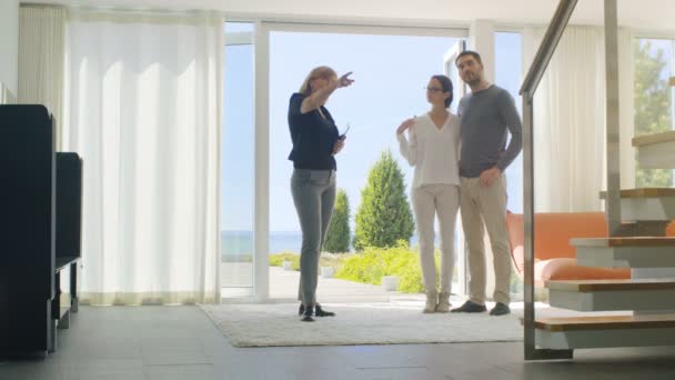 Professioneller Immobilienmakler zeigt stilvolles modernes Haus einem schönen jungen Paar, das auf dem Markt für Kauf / Vermietung eines neuen Eigenheims ist. Haus hat bodentiefe Fenster und Meerblick. — Stockvideo