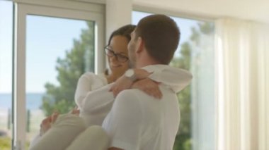 Güneşli oturma odasında, yakışıklı genç Spin-sarılmak onun güzel kız arkadaşı ve onlar kaldırdı. Onlar mutlu ve Sunny Deniz Manzaralı evim.